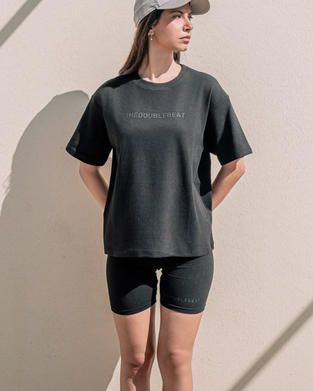 women wearing a dark gray t-shirt and black shorts with 'THEDOUBLEBEAT' branding against a beige wall.