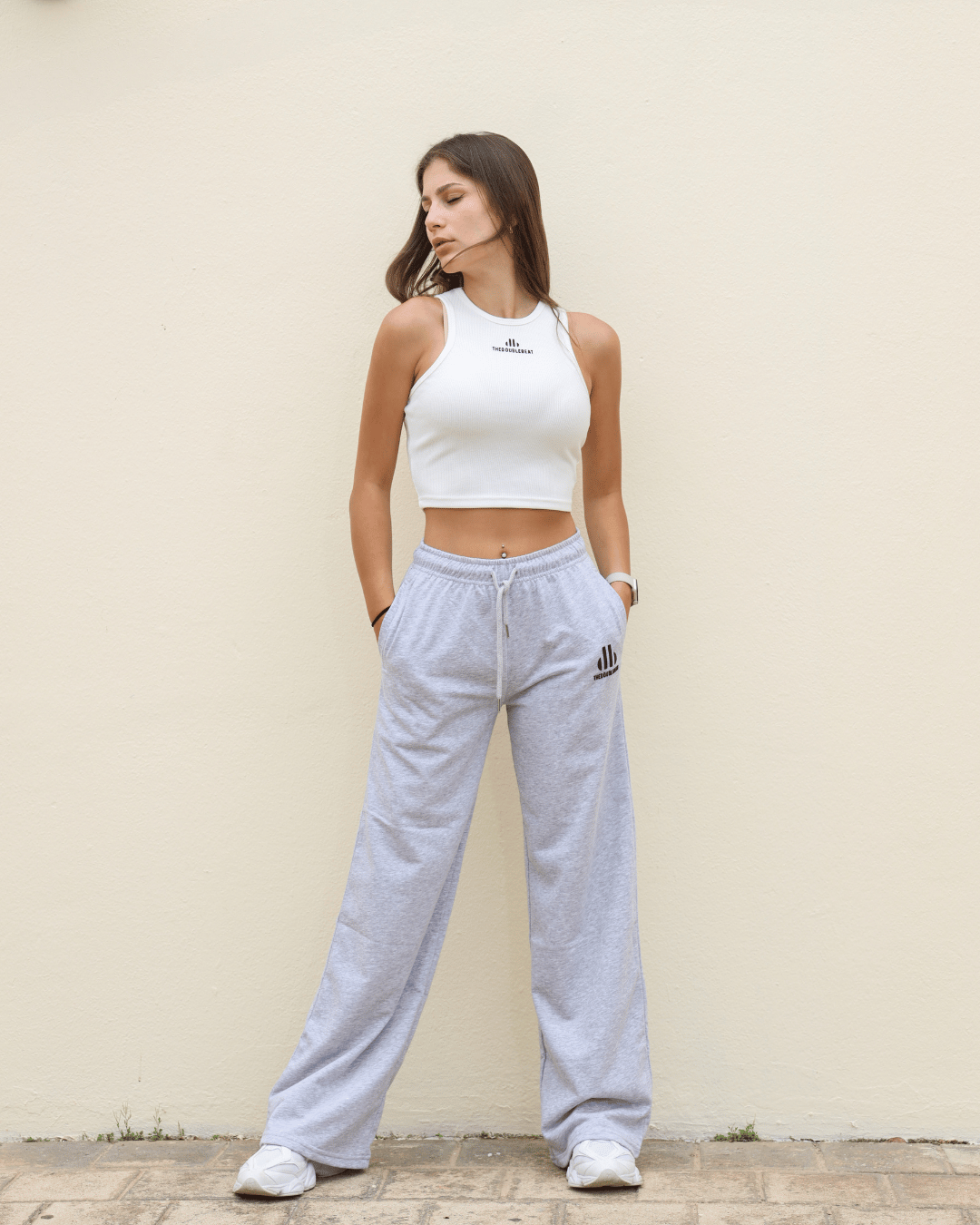 Woman wearing a white crop top and gray sweatpants against a beige wall.