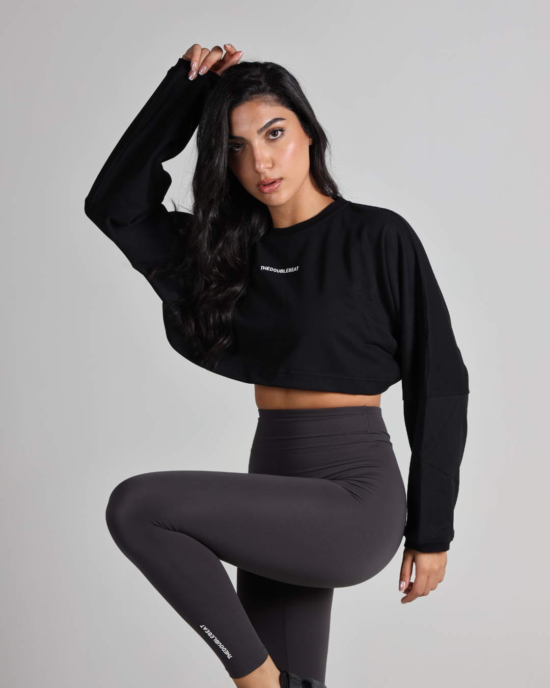 Woman wearing a black long-sleeve crop top and black leggings with a logo on a gray background